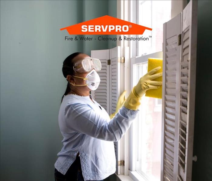A lady with yellow gloves on and a mask cleaning a window with a sponge and the SERVPRO logo at the top.